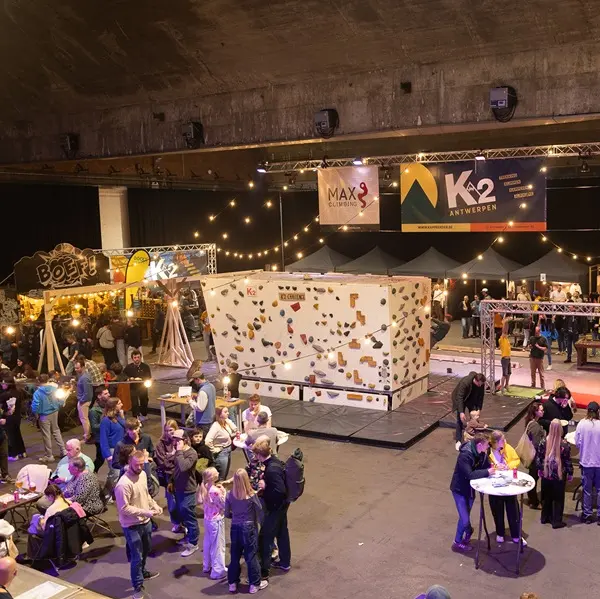 Een evenement binnenshuis waarbij mensen zich rond een centrale witte klimwand hebben verzameld. Op de achtergrond zijn eetkraampjes, lichtslingers en spandoeken te zien, die zorgen voor een levendige en feestelijke sfeer.